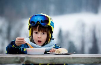 Kind met skihelm en skipak eet soep aan een tafeltje op de skipiste